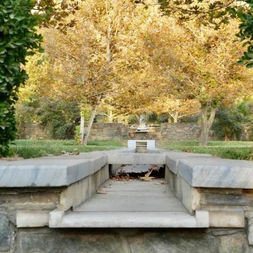 Adobe Express file 3 Central water channel at Bagh-e Babur, part of the Charbagh garden design, surrounded by lush greenery and trees with golden leaves, leading to a fountain in the distance.