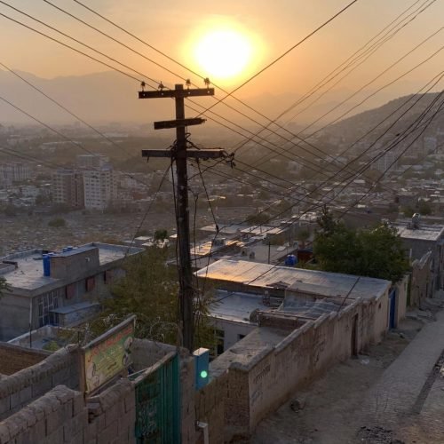 Adobe Express file 3 1 r0tggp1ha2d7f6tc6nngrvdr5uy49mrrm9p8ra06tk Sunset view over Kabul's residential streets, with power lines and distant hills under a hazy sky.