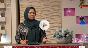 A woman wearing a patterned dress and a teal headscarf speaking on a live television broadcast, standing beside a vase of flowers with a decorative studio backdrop