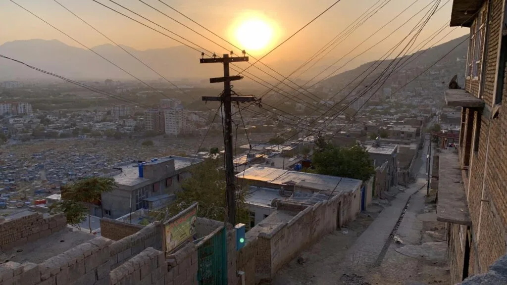 Sunset view over Kabul's residential streets, with power lines and distant hills under a hazy sky.