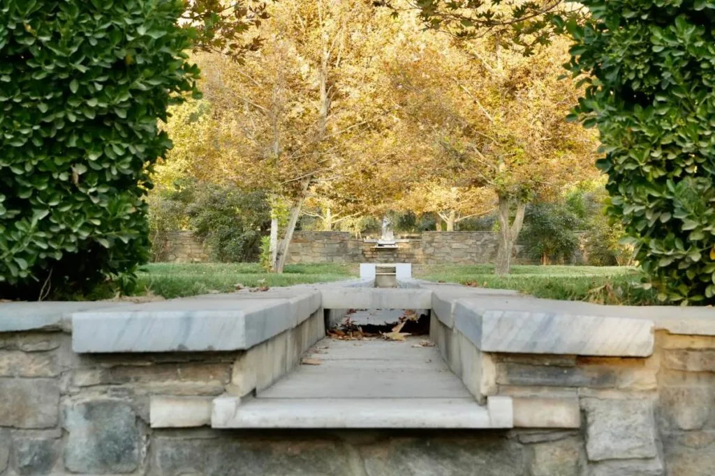 Central water channel at Bagh-e Babur, part of the Charbagh garden design, surrounded by lush greenery and trees with golden leaves, leading to a fountain in the distance.