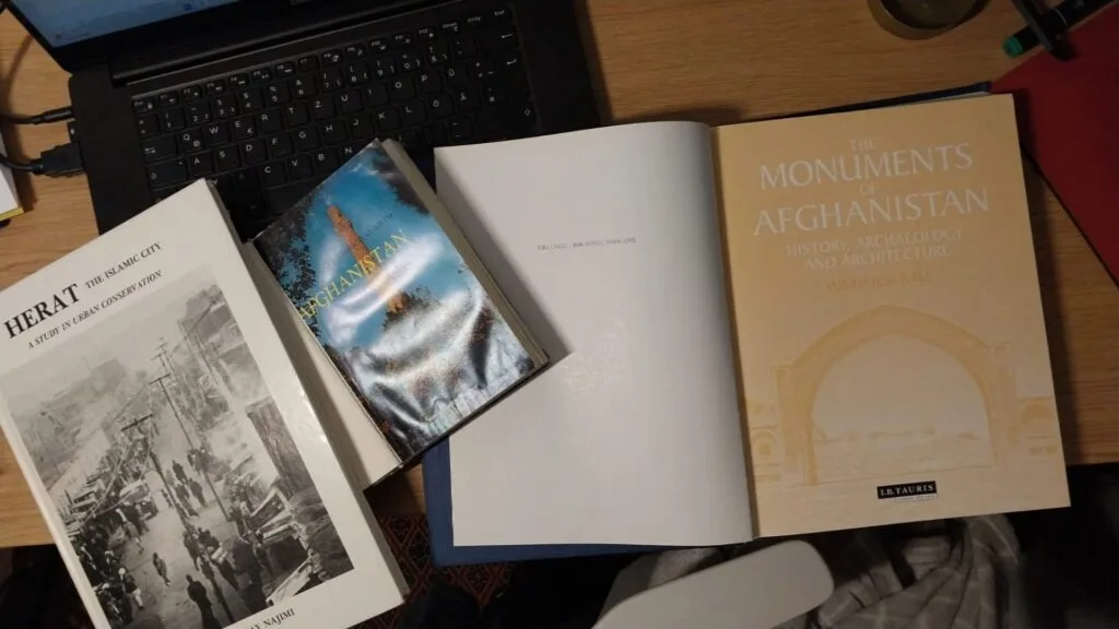 Books on Afghan history and architecture spread on a wooden desk, including titles such as "Herat: The Islamic City" and "The Monuments of Afghanistan.
