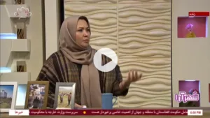 A woman in a headscarf speaking during a live television broadcast, seated at a table with framed photographs and a decorative studio backdrop.