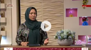 A woman wearing a patterned dress and a teal headscarf speaking on a live television broadcast, standing beside a vase of flowers with a decorative studio backdrop