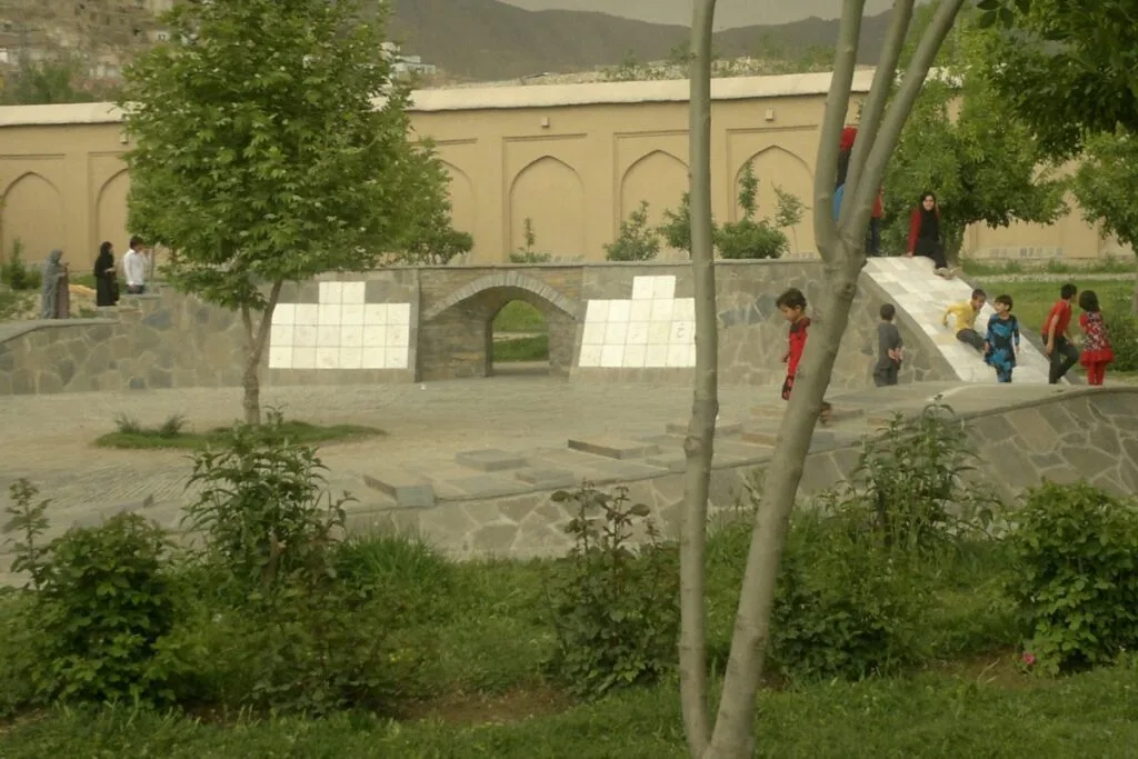 Newly built stone structure with an archway and white tile detailing, part of the modern additions by AKTC to Bagh-e Babur. Children play around the structure, with greenery and trees in the foreground.