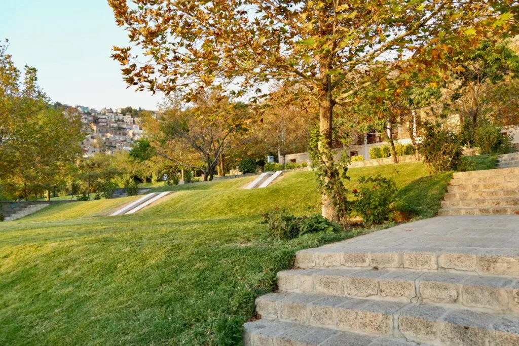 Terraced gardens in Bagh-e Babur, with neatly manicured lawns, stone steps, and autumn-colored trees. Residential homes are visible on the hillside in the background.