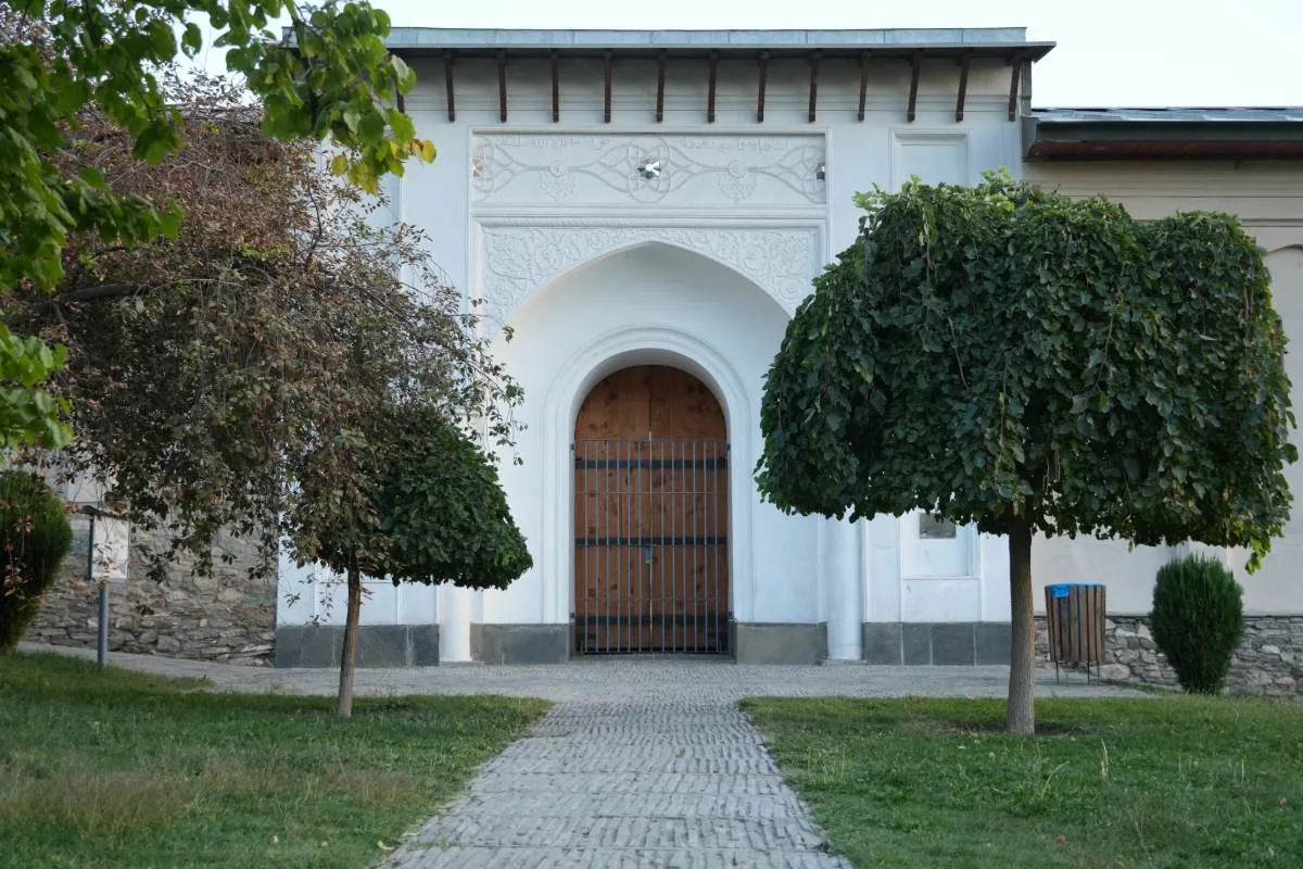 Entrance to the Haram Sarai in Babur's Garden (Bagh-e Babur), featuring a white arched gate with intricate carvings, flanked by manicured trees.