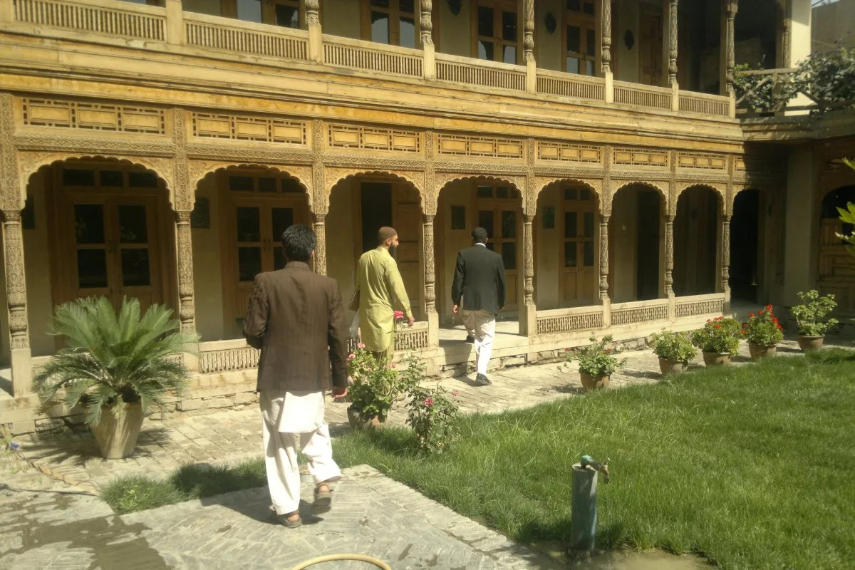 Traditional courtyard house in Afghanistan, featuring intricate wooden carvings and a lush garden with visitors exploring the space.