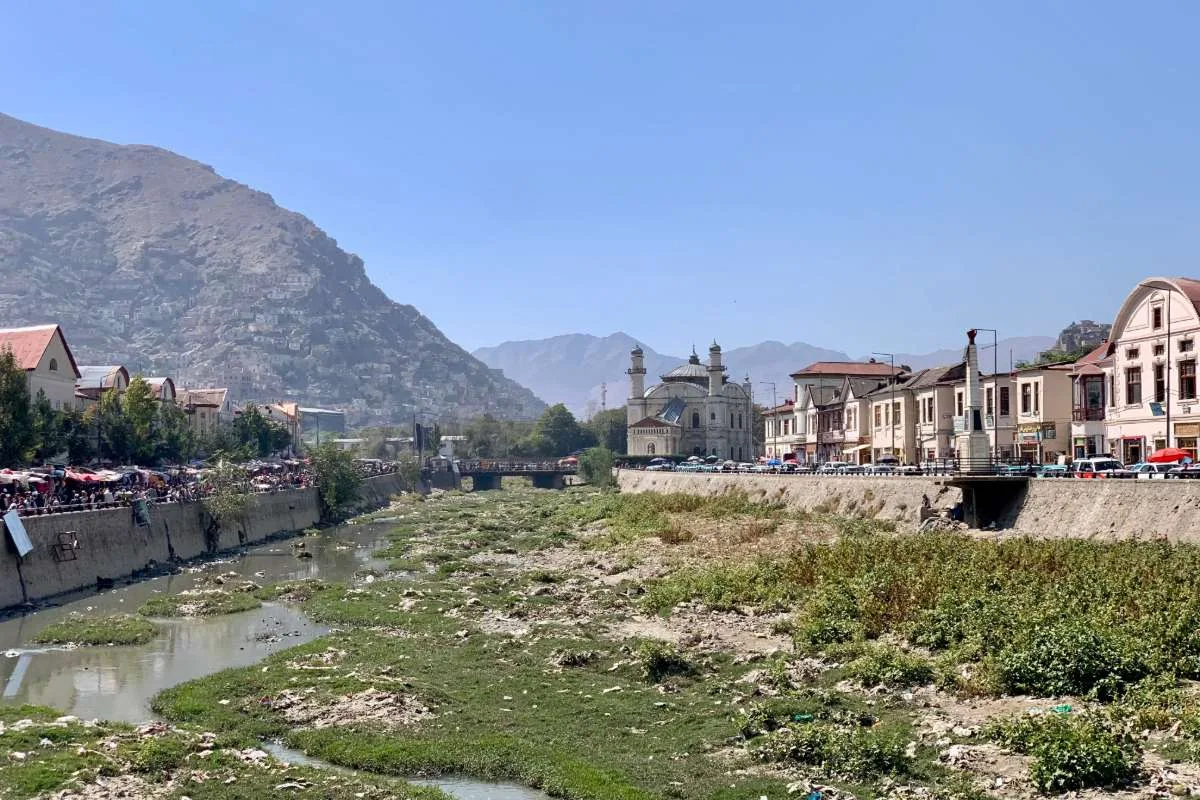 View of Kabul River with the iconic Shah-e-Du-Shamshera Mosque and surrounding hillside homes under a clear blue sky.