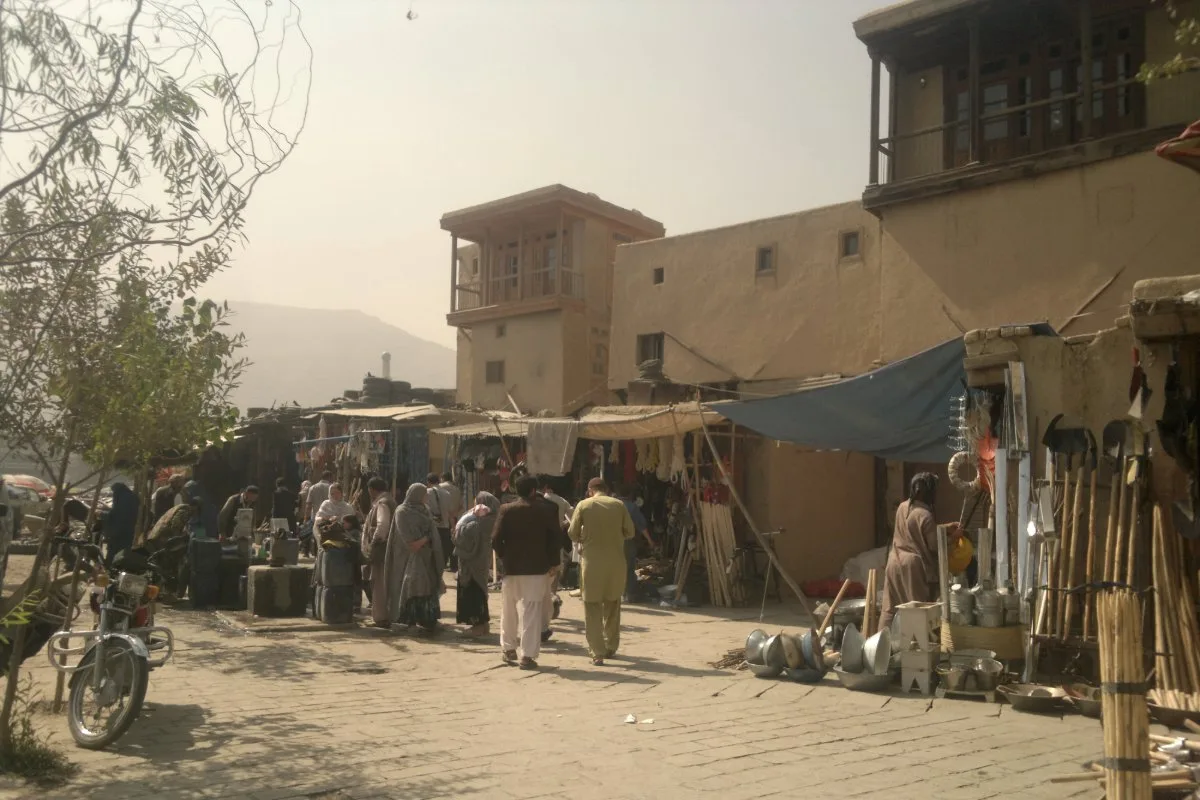 A bustling street scene in the historic Murad Khani district of Kabul, Afghanistan, featuring traditional mud-brick buildings, local vendors, and people shopping under makeshift stalls.