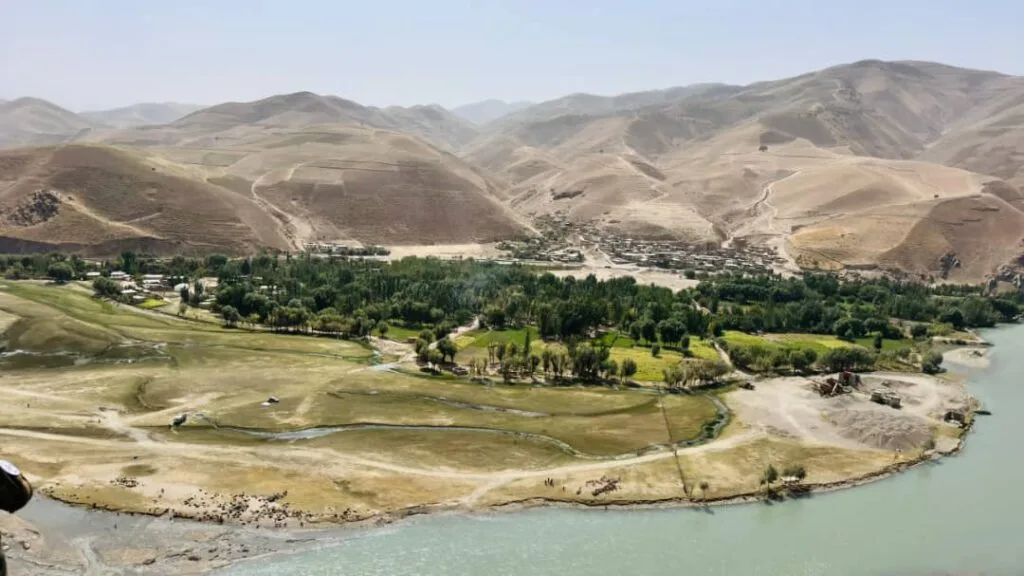 A serene valley landscape in Afghanistan, featuring a lush green area bordered by a river and surrounded by arid mountains under a clear sky