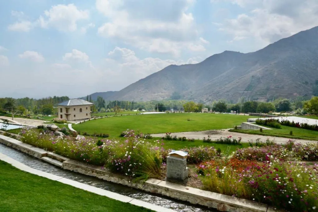 Beautiful garden view at Bala Bagh in Paghman, featuring vibrant flowerbeds, a flowing water channel, lush green lawns, a charming house, and scenic mountain landscapes under a cloudy sky